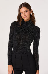 A woman is wearing a long-sleeved, fitted black top with a high, draped neckline. The top is decorated with small, evenly spaced white polka dots throughout. She has paired it with high-waisted black trousers.